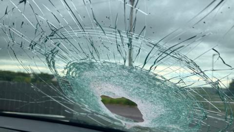 A photo of a smashed up windscreen taken from inside of a car. There is a hole with a lot of damage. The sky is cloudy. A grey fence is in the background. 