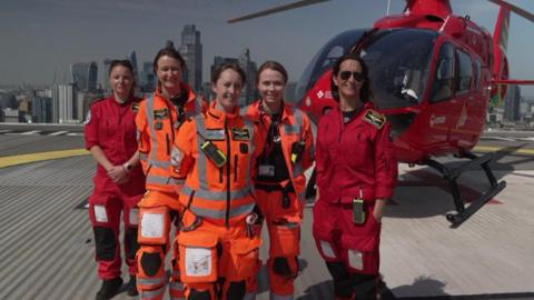 Group photo of the five female crew members on top of a building with the red helicopter behind them and a London skyline in the background