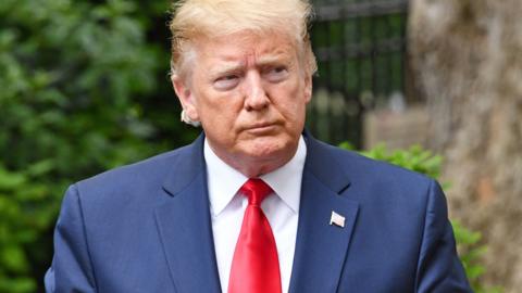 Donald Trump wearing a blue suit, white shirt and red tie, with green foliage visible in the background