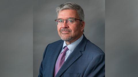 A headshot of Nick Mannion, the Labour leader of Cheshire East Council. He has short grey hair and is wearing clear glasses with a reddish brown frame, a dark blue suit, light blue shirt and purple patterned tie. 