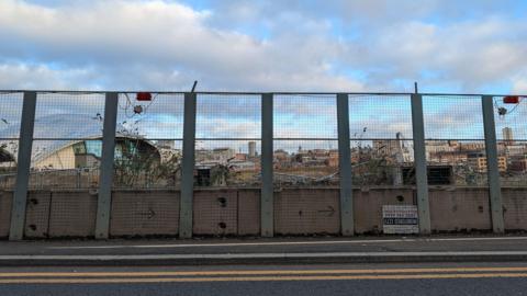 A wire fence with a road in front of it. Double yellow lines are on the edge of the road. Behind the fence on the left is the glass domed roof of the Glasshouse and the top of the white curved roof of the Millennium Bridge can be seen on the right. There are overgrown plants immediately behind the fence which looks aged and run down.