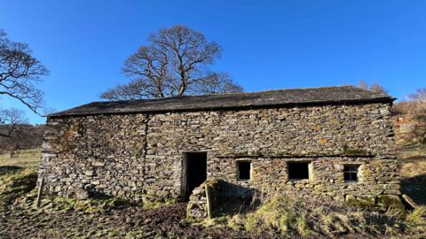 Outside of rural farmhouse with entrance in the middle and three windows to the right hand side. It is in a field on a sunny day. The walls are made of irregularly shaped stones. 