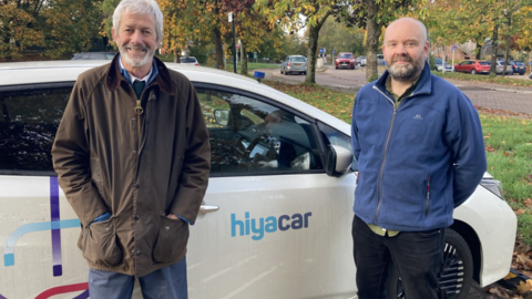 Two men standing beside a white car branded with the logo “hiyacar” in a leafy car park. Autumn trees with orange and green foliage are visible in the background, along with several parked vehicles.