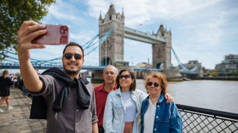 Four tourists standing along River Thames taking selfie with Tower Bridge in the background