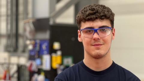 Dane Glenn, a young man with short curly brown hair, wears safety glasses and a blue T-shirt as he stands in the manufacturing area of a wind turbine blade factory in Hull.