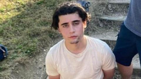 Daniel Khalife, wearing a white t-shirt, is pictured sitting on the ground outside with his hands behind his back after his arrest