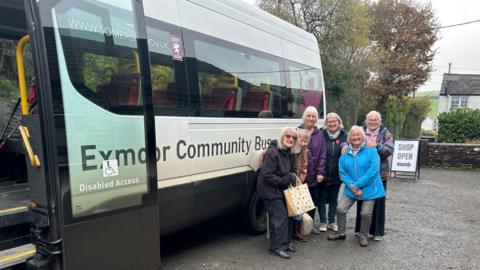 Six ladies posing in front of the minibus that they travel on. They have coats and bags and are smiling. 