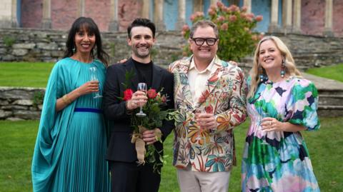 John Cooper wears a black suit and stands holding flowers and a glass of champagne next to Alan Carr. Elegantly-dressed women stand on either side of them, also each holding glasses of champagne. 
