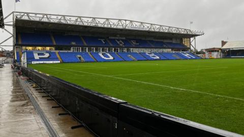 An empty, two-tier stand at Peterborough United's ground. Most of the seats are blue, but there are white sets spelling out the the words "The Posh". ABout half of the green pitch can be seen in front of the stand.