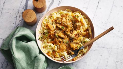 A bowl of vegan mushroom stroganoff sits on a table. Next to the bowl is a pair of wooden salt and pepper shakers, as well as a green tea towel. 