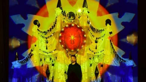 Yellow, red and blue lights are projected onto a huge sculpture of a face. A man dressed in black stands in front of the sculpture.