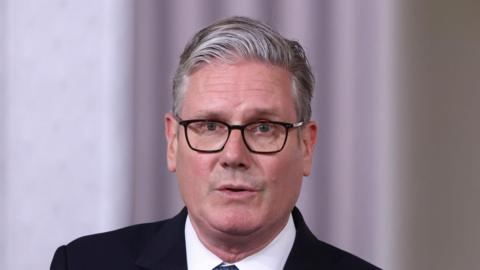 Sir Keir Starmer is looking towards the camera. He has short, grey hair and is wearing a dark suit jacket, a white shirt and a tie.