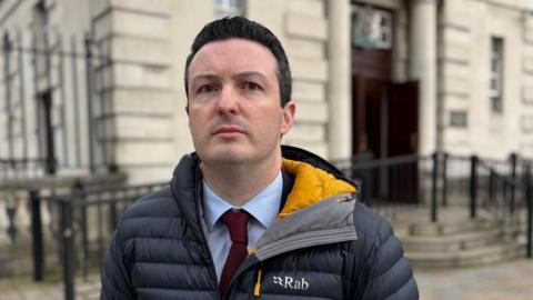 Mr Clayton is looking directly at the camera. He is wearing a navy puffa jacket with the branding RAB on it. He is wearing a maroon tie and light blue shirt.