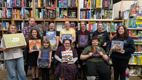 A group of people stand holding board games, smiling. Behind them is a wall of shelves filled with board games.