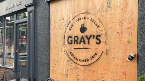 A board covering broken windows with the shop's branding and messages of support written on it.
