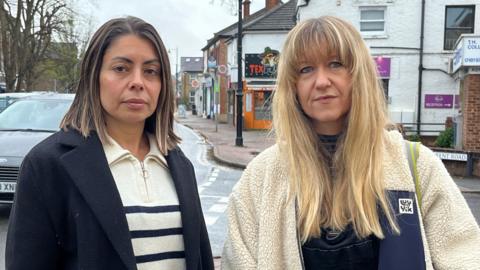 Two women stand next to each other facing the camera, with a busy high street behind them. The woman on the left has dark shoulder length hair and is wearing a cream and blue striped top and a navy jacket, the woman on the right has long blonde hair and is wearing a cream fleece