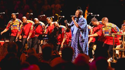 A woman in a blue dress sings in front of a choir of men dressed in colourful red tops and black trousers in a concert hall