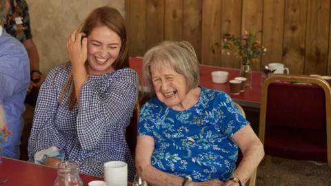 An older and a younger woman laugh as they sit at a table, with hot drinks and water in front of them. 