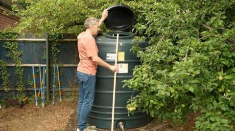 A man is wearing an orange t-shirt and is standing next to a tank of oil.