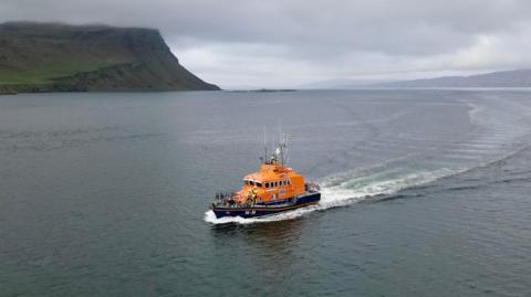 An orange and black coloured lifeboat is powering through the sea off the coast of Skye. White disturbed water can be seen in the wake of the boat.  In the distance is a rocky stretch of coastline partly obscured by low cloud.
