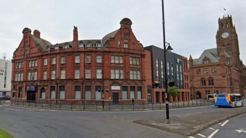 The Majestic Hotel in its current form. It is a red-brick, four-storey building with large windows on the corner of a road. Its gables on either side look like rounded triangles. There are short black safety railings in front of it. In the background is a church clock tower and a bus is going past.