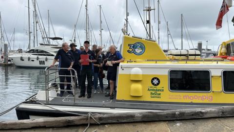A group of men holding notebooks standing on the back of the Flying Christine III marine ambulance which is docked in a harbour with grey skies behind.