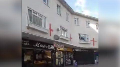 A row of shops is shown on the ground level with flats spread across the two floors above them, with the red flag of St George painted on the white walls of the homes.