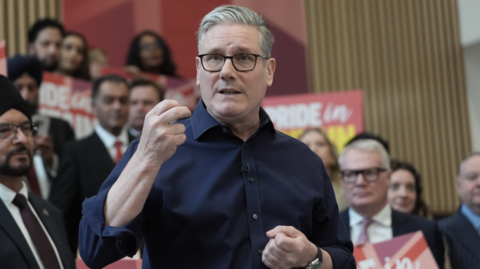 Keir Starmer wearing a blue shirt, standing in front of a group of people, some of whom are holding Labour placards