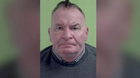 A police mugshot of Mark Thone, a man with receding grey and black hair. He is wearing a dark grey jumper with a checked shirt underneath. His mouth is turned down at the corners and one eyebrow is raised as he looks directly at the camera.