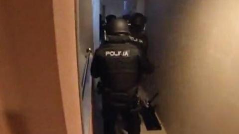 Armed police storm an apartment. They are wearing black clothing with Policia written on it
