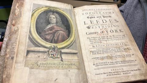 An historical open book on display. On the left page is a coloured portrait of a man with long, curled hair, dressed in early 18th‑century clothing. Printed text identifies him as  Radulphus Thoresby, Leodiensis, S.R.S. 1712. The right page shows the book’s title page, filled with  ornate text. The title reads 'Ducatus Leodiensis'.