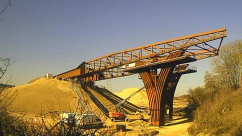 Bridge slowly being put into place on a sunny day, workers and vans can be seen below it.