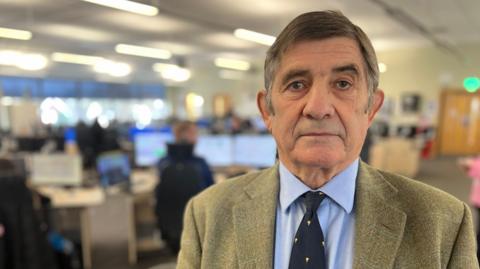 Man with smart brown and grey hair in a side parting looks to the camera. He is right of centre with a call centre in the background. He is wearing a brown suit with a blue tie and shirt.