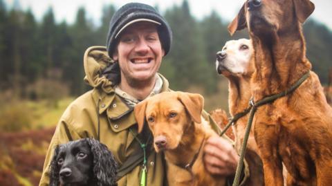 Sam Cropper in the centre of the image. He is smiling at the camera. He is sitting down and wearing a khaki jacket and a dark-coloured hat. A small black dog is sitting in front of him. He is cuddling a light brown dog on his left side. A dark brown dog is looking up in the foreground and a light-brown dog is behind it looking off camera. All are sitting in front of a row of trees.