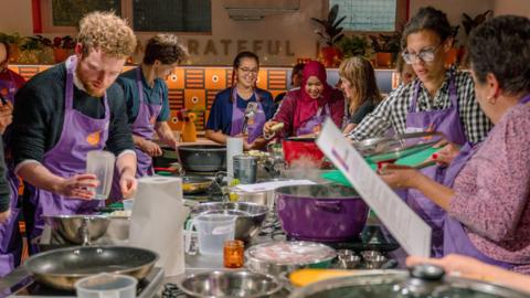A big group of people from multiple different nationalities are standing around a professional kitchen table, stirring, pouring and chopping food. They are all wearing purple aprons. Many people are smiling, while others look very focused. There are lots of different kitchen utensils on the table, knives, saucepans, frying pans, bowls and kitchen roll. 
