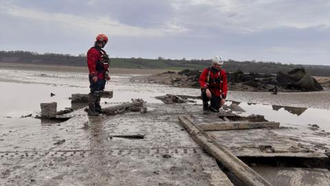 Two people wearing red safety gear and helmets standing in wet muddy sand looking at bridge remnants covered in silt. It is an overcast day.