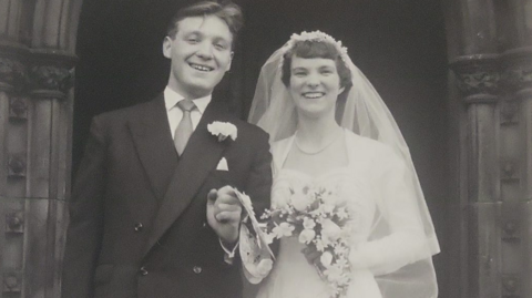 A young John and Barbara are standing together in a black and white image on their wedding day. Barbara is wearing a white wedding dress and holding flowers and John is dressed in a formal suit and holding her hand. They are both smiling at the camera.