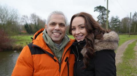 Nick Samiotis with his daughter Tabitha in front of their garden in Saham Toney in Watton. Nick is wearing a bright orange duck down coat with a khaki hoodie underneath and is smiling at the camera. His daughter, Tabitha is wearing a black coat with a fur trimmed hood and a belt. There is a duck pond in the background and there is an overgrown gravel driveway peeling off behind them in the distance through some trees.