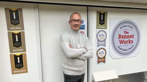 A man standing in front of a white outbuilding with awards signs on it. A sign says "The Rennet Works - hand made cheese". 