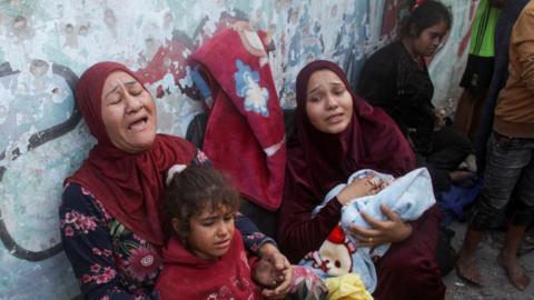 Two women cry out as they lean against a wall and hold two children - one a young girl and the other a baby wrapped in a blanket - in a school sheltering displaced people in Gaza. Another young woman is seen in the background.