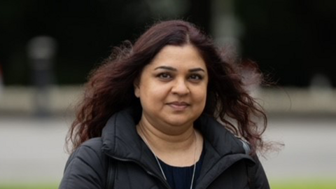 Tanya Nasir has reddish-brown long and slightly curly hair, she has dark brown eyes and is wearing a silver necklace and light black puffer jacket