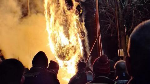 A number of silhouetted heads can be seen standing in front of a burning effigy