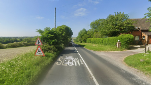 Google maps street view of the A346 in Ogbourne St George, Wiltshire. The road is straight with "Slow" painted on it in white capital letters. Two triangle shaped road signs can be seen on the left of the road - a junction sign and a slippery road sign. A house can be seen on the right. 