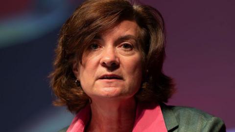 Eluned Morganin front of a partially red and blue background, wearing a green jacket and a red shirt.