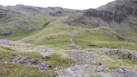 Looking back down on Sty Head while ascending Great Gable by the Breast Route. The path to Sprinkling Tarn is ahead centre. There is a rocky path leading up to a crag which is covered in green grass.
