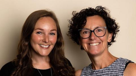 Two women. One with long dark, brown hair and one with short curly brown hair and glasses. They are both smiling at the camera.
