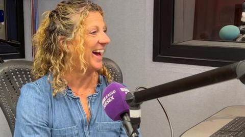 Olympian Sally Gunnell, wearing a light blue denim shirt, sits in the BBC radio studios at Brighton, speaking into a microphone with a purple branded windsock cover.