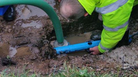 A man in a bright yellow jacket leans down as he installs equipment in a muddy trench