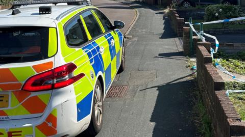 A close-cropped image of the scene outside the house police were called to. A police car takes up one half of the screen, parked on the kerb, photographed from behind. Blue and white police cordon tape can be seen around the entrance to one front garden. 