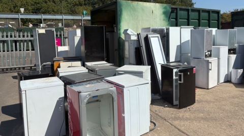 More than a dozen, mainly white, appliances are next to each other outside. Railings and trees are in the background.